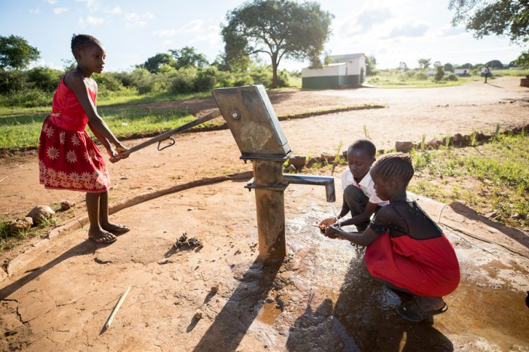 Two girls and a boy collect water from a hand pump